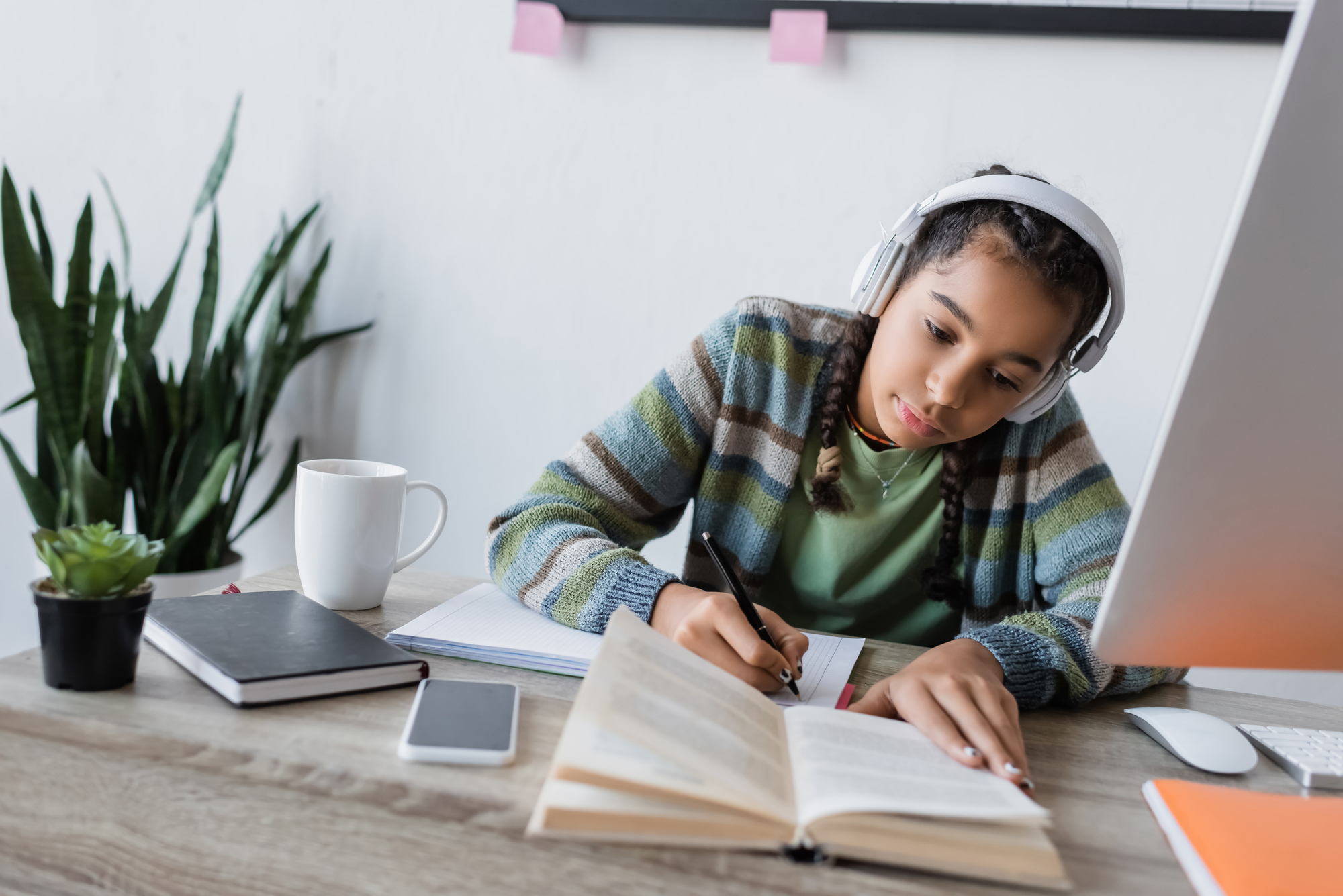 Girl studying on an online GCSE course for homeschooling, sitting at a desk, wearing headphones and writing in a book